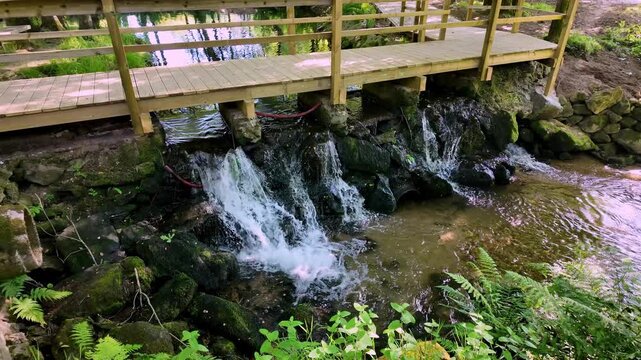 Scenic view of a wooden pedestrian bridge over a small cascading waterfall in the lush green environment of Parque de Lazer de Nadais under bright natural light.