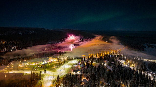 Aerial view of Luosto ski resort at night with fireworks exploding over snow-covered forests and the aurora borealis in the sky Sodankyla, Lapland, Finland.