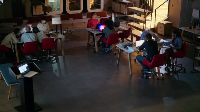 A diverse business team collaborates around tables in a modern office environment. Professionals are engaged in discussion and working on laptops during a productive meeting.