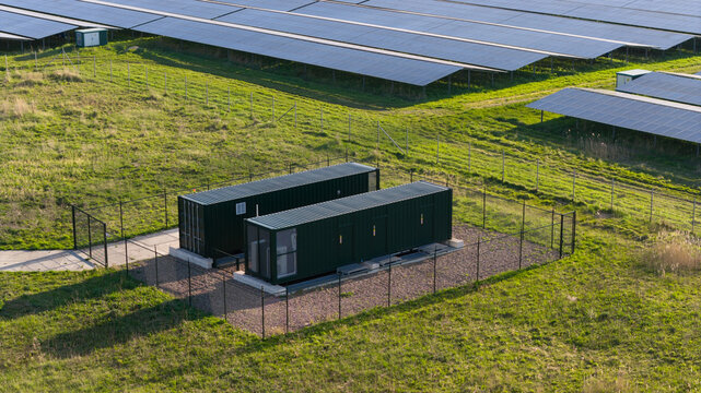 Aerial view of a solar farm featuring rows of photovoltaic panels and green battery storage containers within a fenced enclosure in Farmsum, Groningen, Netherlands.