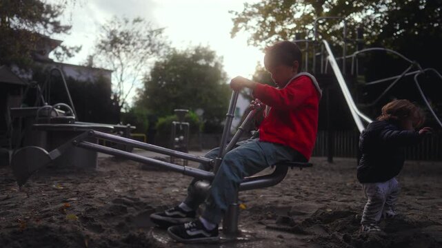 Older boy operating playground sand digger with focused effort while toddler stands beside exploring sand creating authentic outdoor moment showing parallel play curiosity and natural childhood rhythm