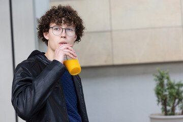Teen male standing by tan wall sipping orange juice through straw from cup wearing jacket, glasses © wavebreak3