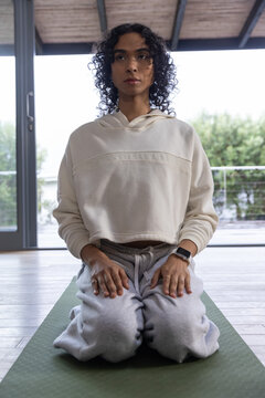 Non-binary person kneeling on green mat, wearing hoodie, showing smartwatch and rings by windows