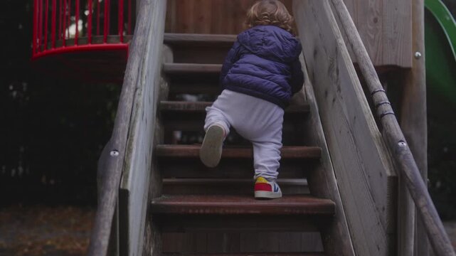 Toddler climbing wooden playground stairs with determination during outdoor play creating natural moment showing childhood exploration early independence movement and gentle focus in cool weather