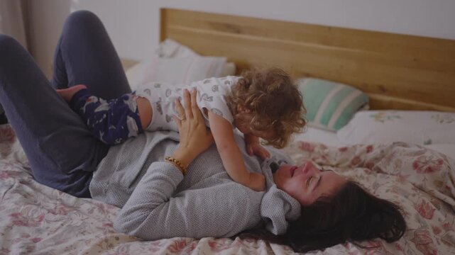 Mother playing with toddler on bed in joyful morning moment as they laugh cuddle lift and lean toward each other creating warm affectionate playful connection full of movement
