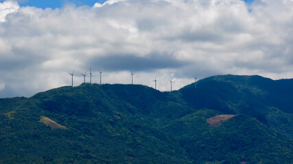 Wind turbines on a mountain ridge. Renewable energy infrastructure stands above lush tropical hills beneath dramatic clouds and changing weather conditions. © Houston