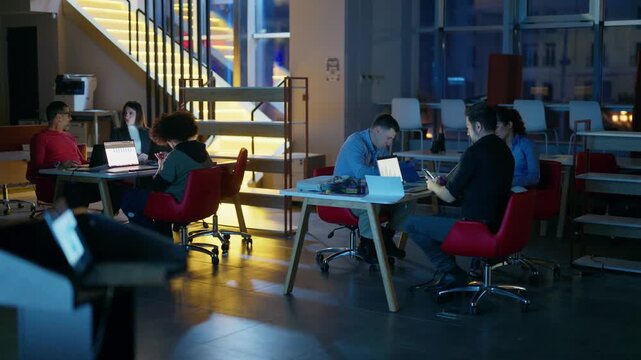 A diverse team of professionals works collaboratively on laptops in a contemporary office setting late at night.