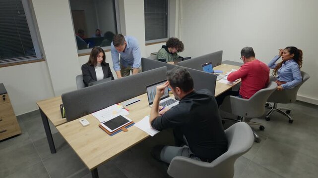 Employees are diligently working on computers and discussing projects in a modern office. Businesspeople are busy with their daily tasks at their individual workstations.