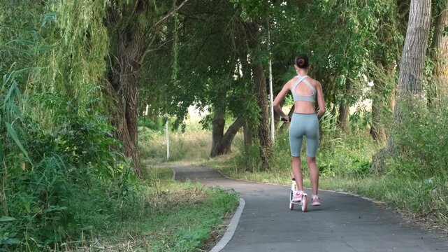 Back view of young girl athlete riding white kick scooter through green summer park. Concept of joy and dynamic lifestyle