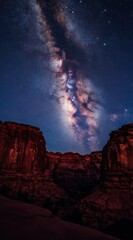 Fototapeta premium Vast Milky Way galaxy arches over dramatic red rock canyon at night. Cosmic dust clouds and stars create spectacular celestial display. Dark landscape below shows rugged desert formations.
