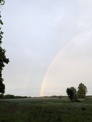 Fototapeta premium A breathtaking rainbow arches across a vast, overcast sky above a tranquil rural landscape, with fields stretching towards the horizon
