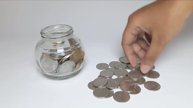 Closeup of a hand placing Indonesian rupiah coins into a glass jar indoors on a clean white background for saving, budgeting, investment, and personal finance concepts.