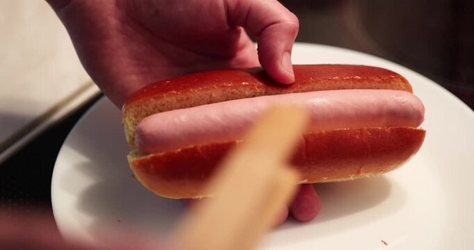 A person places a hot dog in a bun while adds ketchup and put it on a white plate in the kitchen.