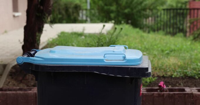A person opens a black trash bin beside a building to dispose some pizza boxes after lunch.