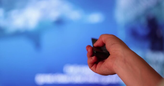 Man hand holding television remote control pointed toward LED screen to switch channels in home environment.