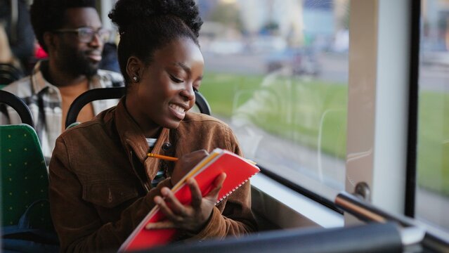 African American woman holding red notebook and pencil while sitting on bus seat and looking out window. Passenger writing and thinking about words while working or studying during commute.