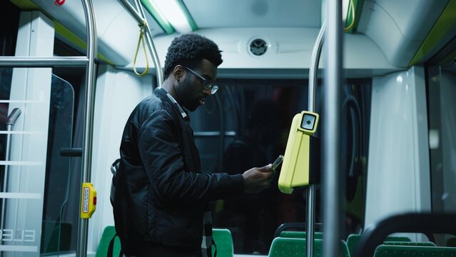 African American man wearing glasses and black jacket holding smartphone near validator. Male using contactless payment while standing inside city bus at night. Modern public transport technology.