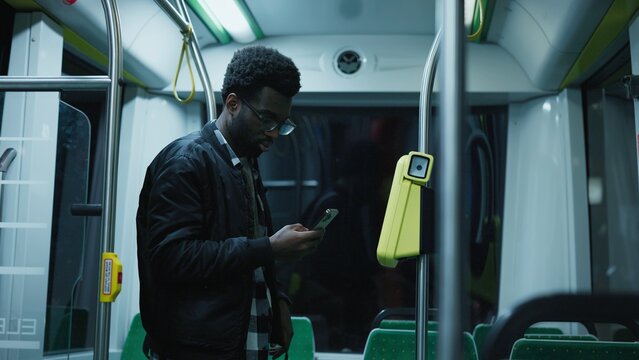 African American man wearing glasses and black jacket holding smartphone near validator. Male using contactless payment while standing inside city bus at night. Modern public transport technology.