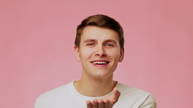A young man with light skin is sending a kiss gesture towards the camera. He has short brown hair and is wearing a white shirt. The background is a soft pink color.