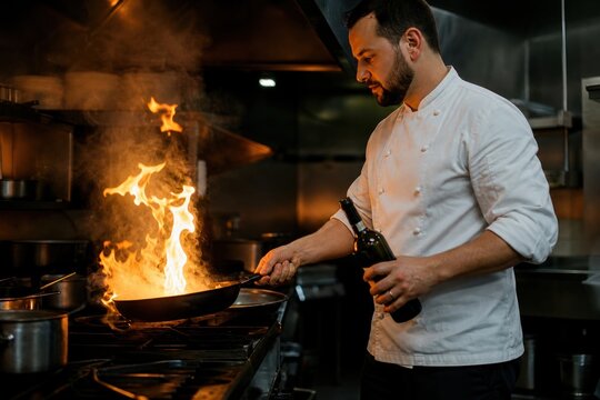 Professional chef performing flamb&eacute; technique in modern kitchen, dramatic orange flames igniting wine over hot pan, intense culinary action, high-end restaurant cooking atmosphere