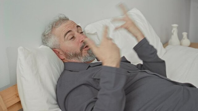 Middle aged man gesturing with hands while reclining on a bed in a building bedroom; exasperation release.