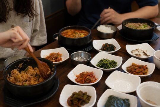 Traditional Korean family-style meal with diverse banchan side dishes, rice, stew, and kimchi served on wooden table in cozy restaurant setting, vibrant colors, communal dining experience