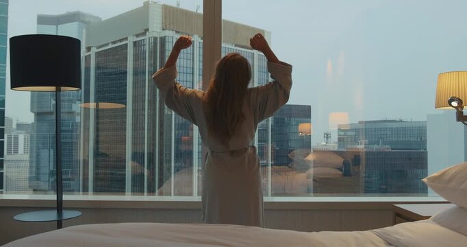 Woman in bathrobe stretching arms while looking at urban city skyline from high-rise hotel room window during morning. Back view of female guest enjoying travel and city view.