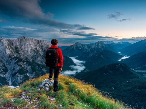 Hiker with backpack standing on a grassy mountain ridge enjoying the freedom of exploration overlooking a dramatic alpine landscape at sunset, Mangart, Julian Alps, Italy-Slovenia border.