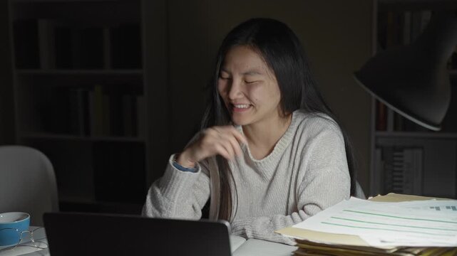 Woman studying at laptop holding a pen and smiling with eyes closed among a stack of papers and a desk lamp in building; contentment.