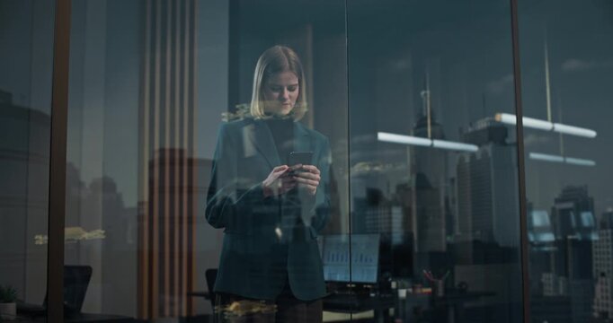 Businesswoman Using Smartphone App Behind Interconnected Glass Office Window Overlooking Futuristic Smart City With Autonomous Delivery Drones Flying Past Skyscrapers at Dusk.