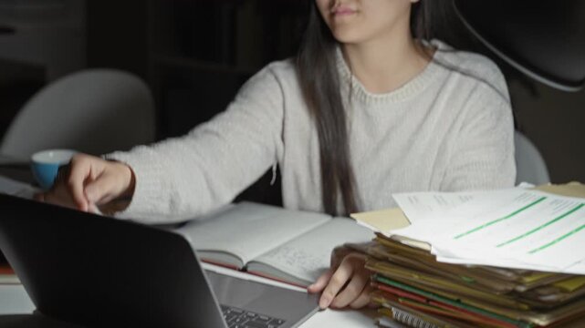 Woman studying at laptop with papers and glasses on table, eyes closed in brief rest, stack of folders visible in a building; tired concentration night.