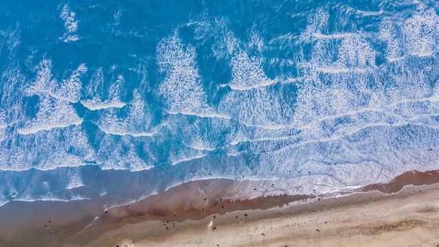 Vista a&eacute;rea cenital de olas rompiendo en una playa de arena con personas lejanas