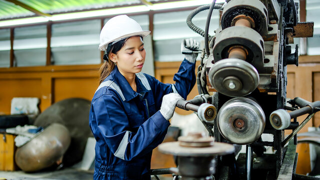 Asian female technician in blue uniform and hard hat operating heavy machinery in industrial factory, focused professional woman worker at metal manufacturing plant production line.