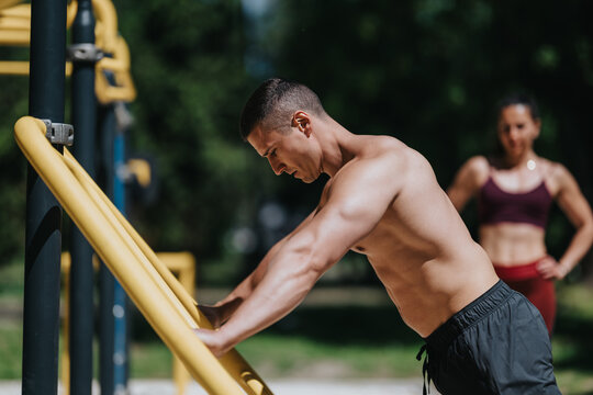 Shirtless fit man leans on yellow parallel bars performing a bodyweight exercise. A female trainer stands in the background observing the outdoor workout.