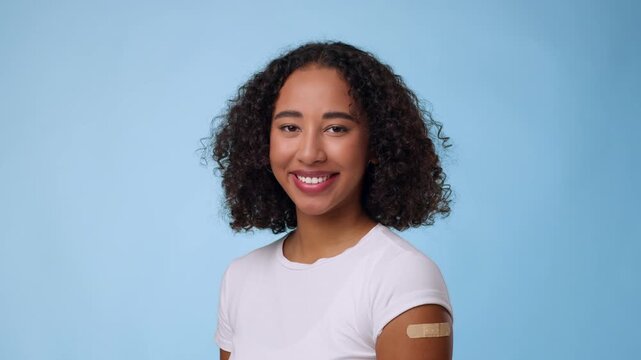 An African American woman with curly hair stands smiling in front of a light blue background. She wears a white shirt and has a bandage on her arm, suggesting she has just received a vaccination.