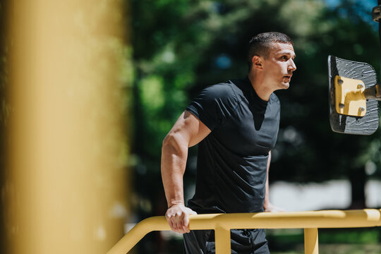 A fit man performs dips on outdoor parallel bars during a daytime workout. He trains upper body strength on park equipment in a focused outdoor exercise session.