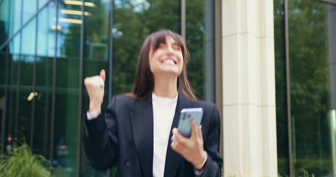 Close-up of an amazed businesswoman holding a smartphone, happily reacting to a notification and expressing excitement through gestures outdoors. Positive emotions and pleasant feelings concept.