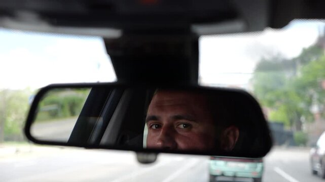 Eyes of male smiling driver reflecting in rearview mirror while riding motor auto. Young man looking concentrated to urban road during operating vehicle. Confident guy driving modern car at city