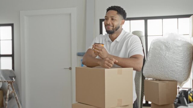 Man holding coffee cup and leaning on stacked cardboard boxes, relaxed smile while surrounded by bubble wrap and packing supplies in a building; new beginning contentment.