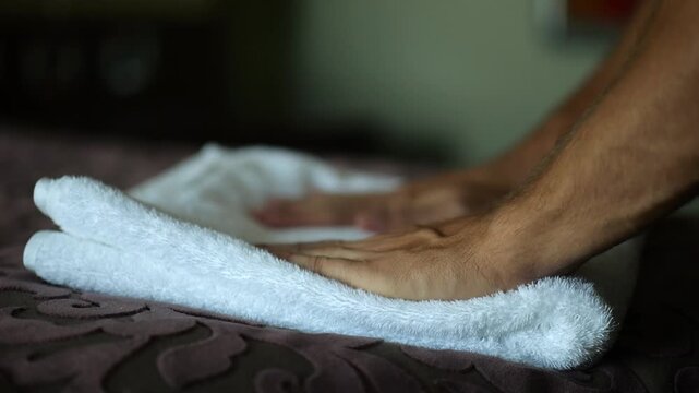 Male hands of housekeeping adjusting soft white terry towel on a neatly made bed in a modern hotel room. Service personnel preparing clean bedding and hospitality interior for guest accommodation