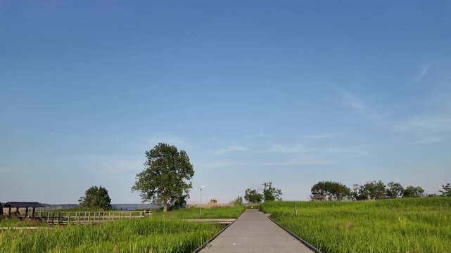 POV walking on a wooden boardwalk towards a traditional wooden water wheel in Kasumigaura Sogo Park under a clear blue sky