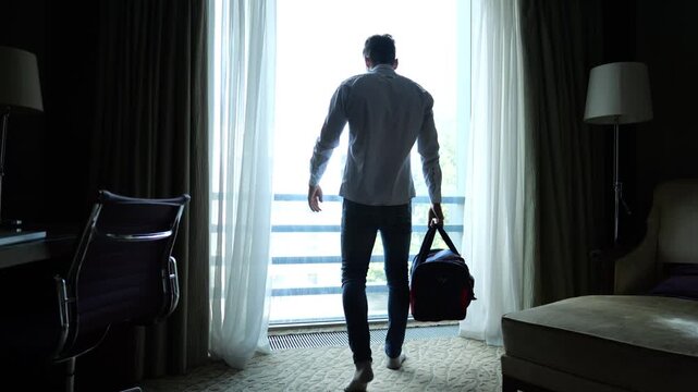Tired young businessman with travel bag standing near large window in modern hotel room after long trip. Exhausted man leaning on glass and resting after business travel showing fatigue and stress