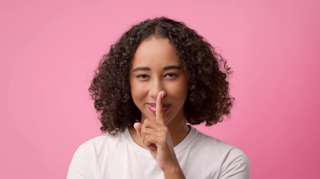 A young woman with curly hair is playfully holding her finger to her lips against a pink background. She appears engaging and cheerful while encouraging quietness or secrecy.