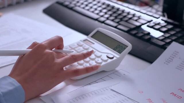 Focused professional hand pressing calculator button holding pen over business document next to computer keyboard on office desk for finance tax