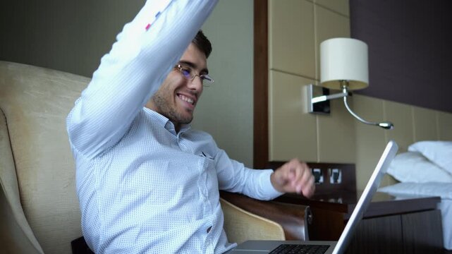 Young businessman working on laptop in modern hotel room and reacting to good news with excitement and happiness. Happy man celebrating success while using computer show achievement and motivation