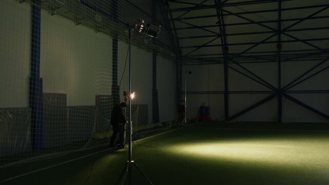 Wide shot of two Caucasian male video production crew technicians installing tripods with professional lighting equipment at indoor sports arena for filming footage for documentary or commercial