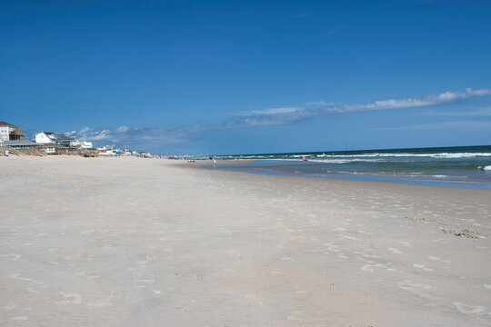 A wide sandy beach along the Outer Banks in North Carolina with gentle ocean waves under a clear blue sky. Elevated beachfront houses line the left side of the shoreline, several people are visible. 