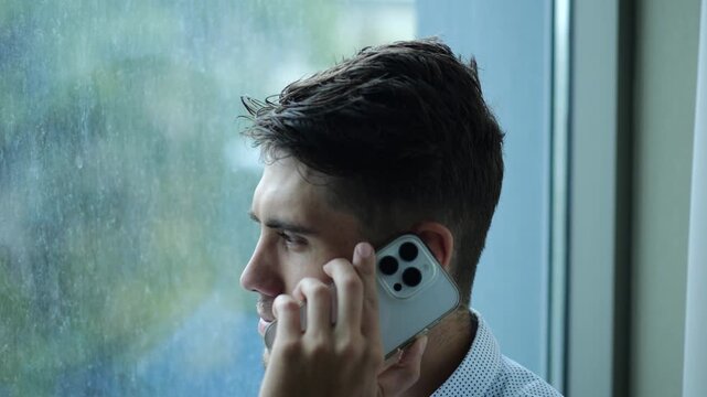 Portrait of male entrepreneur in shirt speaking by smartphone near large window into skyscraper. Young confident businessman talk on phone at office. Handsome manager having conversation on cellphone