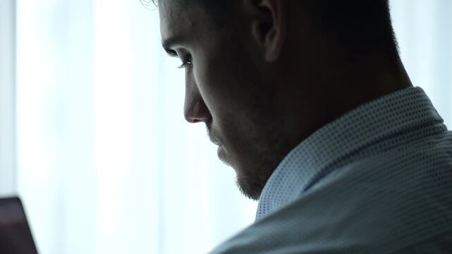 Close up portrait of young entrepreneur standing near large window in modern office and working on notebook. Handsome businessman in shirt using notebook for job with bright day light at background