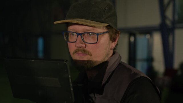 Medium close-up of face of professional Caucasian male videographer in baseball cap and glasses adjusting camera frame while preparing to shoot at sports arena, giving gesture directions to crew